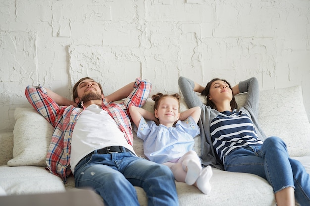 Photo relaxed young family resting and dreaming about new home on comfortable sofa together at home, happy young parents with little daughter relax enjoying nap relaxing or meditating.