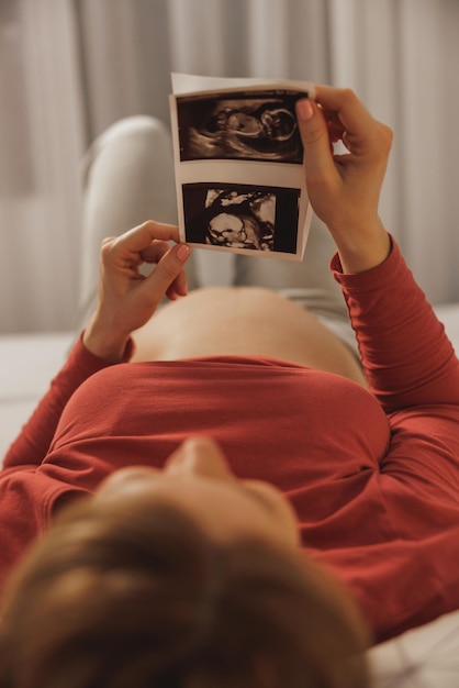 Photo relaxed young future mother looking at ultrasound of her baby while relaxing on a bed in bedroom.