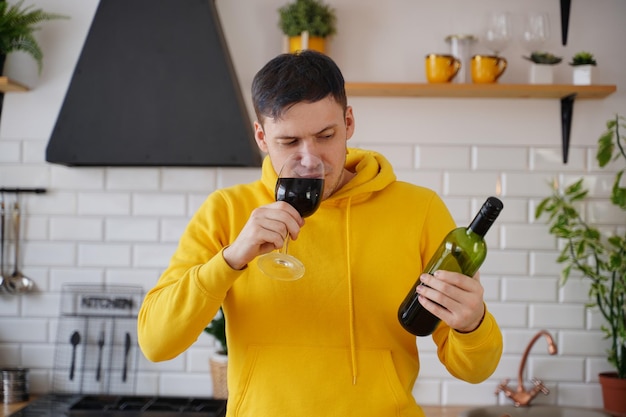 Relaxed young man drinks red wine and looks on bottle Adult guy resting with alcohol in kitchen