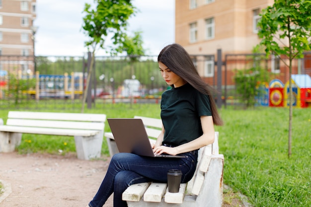 Relaxed young woman in a green T-shirt and with a backpack sitting on a wooden bench drinking coffee and browsing on her laptop