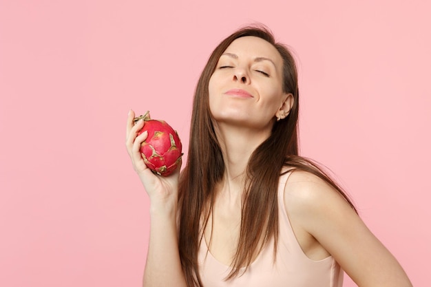 Relaxed young woman keeping eyes closed holding fresh ripe pitahaya, dragon fruit isolated on pink pastel wall background in studio. People vivid lifestyle, relax vacation concept. Mock up copy space.