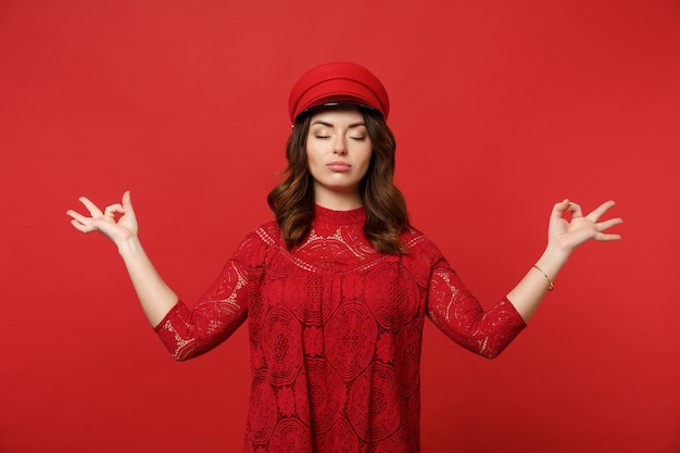 Relaxed young woman in lace dress, cap keeping eyes closed, hold hands in yoga gesture, relaxing meditating isolated on red background. People sincere emotions, lifestyle concept. Mock up copy space.