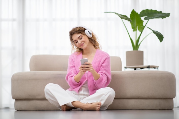Relaxed young woman listening music in headphones and using smartphone at home