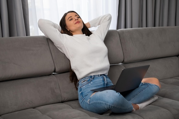 Relaxed young woman lounges on comfortable sofa arms crossed behind head in cozy modern living room