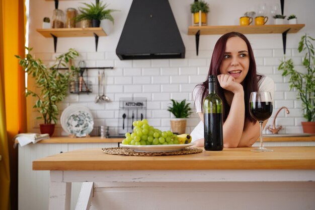 Расслабленная молодая женщина позирует стоя за кухонным столом Взрослая брюнетка отдыхает с алкоголем и фруктами на кухне
