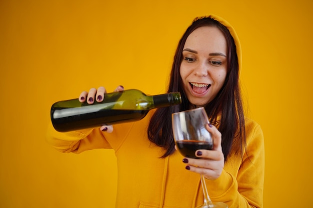 Relaxed young woman pours red wine into glass on yellow background Adult brunette fills transparent glass with alcohol