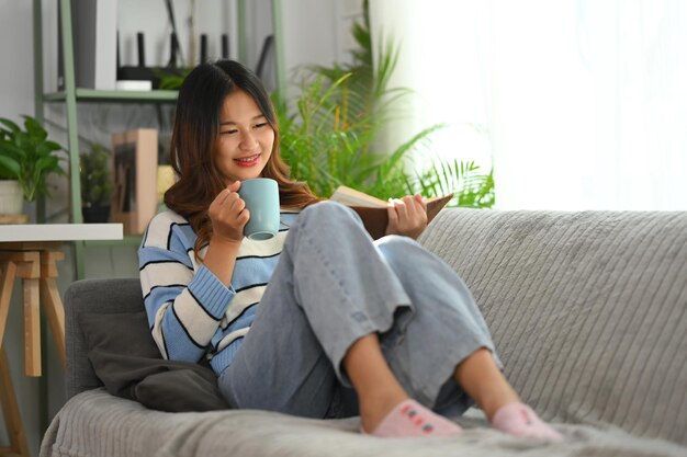 Photo relaxed young woman reading book and drinking coffee on couch at home
