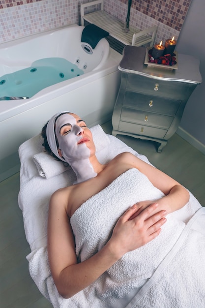 Relaxed young woman resting lying with clay facial mask in spa. Medicine, healthcare and beauty concept.