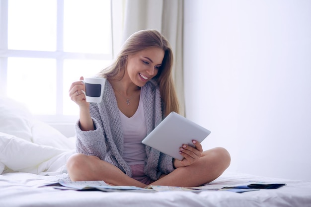 Relaxed young woman sitting on bed with a cup of coffee and digital tablet