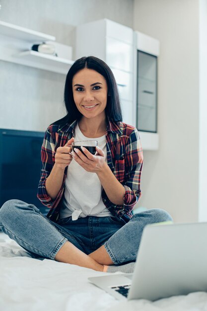 Relaxed young woman sitting cross-legged on her bed and smiling while holding a cup of coffee