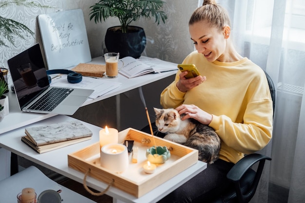 Relaxed young woman with cat sitting near table with burning candles and aroma sticks at home using