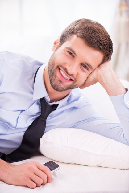 Relaxing after day working. Handsome young man in shirt and tie lying in bed and smiling