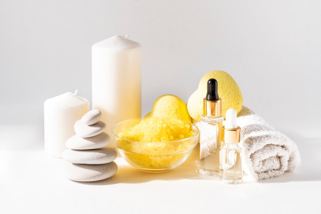 Relaxing bath products composition with yellow heart shaped bath bombs, sea salt scrab, body serum, candles, towel and stone stack on a light wall. Relaxation, spa and body treatment concept.