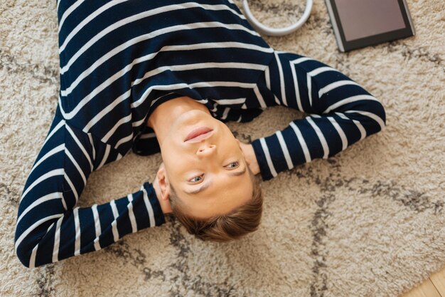 Relaxing on floor. Handsome concentrated fair-haired well-built young man lying on the floor and thinking and his tablet lying near him