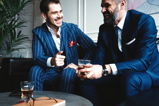 Relaxing. Full length of two young handsome men in suits holding glasses and looking at each other while resting indoors.