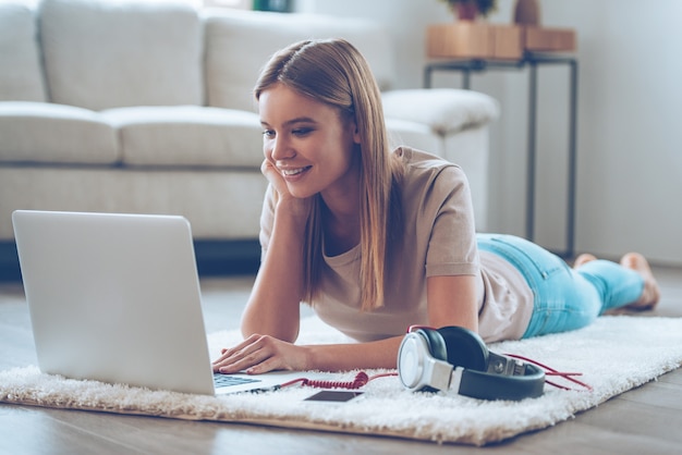 Relaxing at home. Beautiful young woman using her laptop while lying on carpet at home