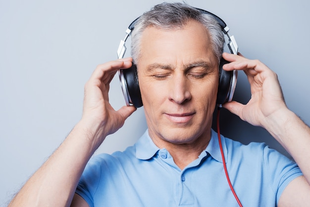 Relaxing music time. Portrait of senior man in headphones listening to music keeping eyes closed while standing against grey background