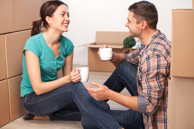 Relaxing in new house. Cheerful young couple sitting on the floor and drinking coffee while cardboard boxes laying all around them