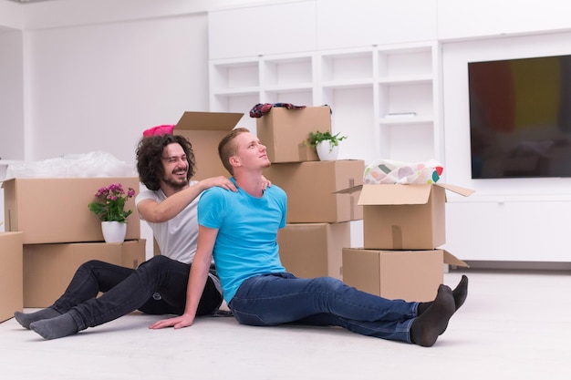 Relaxing in new house. Cheerful young gay couple sitting on the floor while cardboard boxes laying all around them