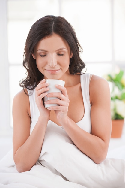 Расслабляйтесь с чашкой свежего кофе. Привлекательная молодая улыбающаяся женщина, сидящая в постели и держащая чашку кофе