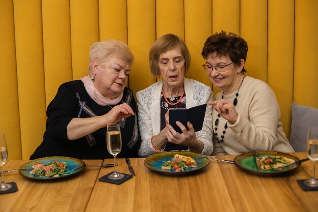 Relaxing women dressed in white, beige and black looking at phone, having pizza on plates, dicussing photos while celebrating their meeting or holiday in modern cafe or restaurant with orange walls