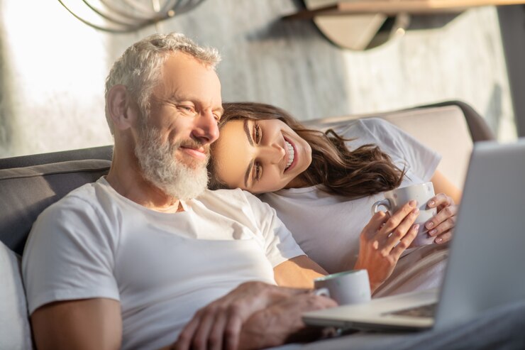 Photo reliable shoulder. happy young woman touching head on shoulder of gray-haired smiling man looking at laptop in bed