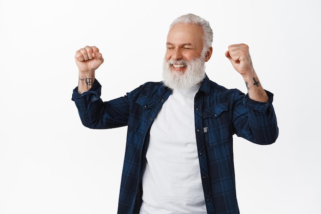 Relieved and excited old man winning prize raising fists up and say yes smiling satisfied achieve goal success celebrating victory win standing over white background