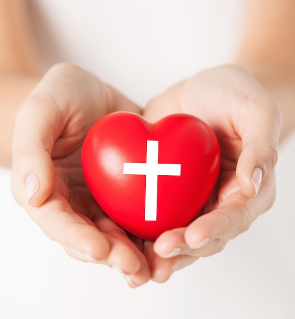 Religion, christianity and charity concept - female hands\
holding red heart with christian cross symbol