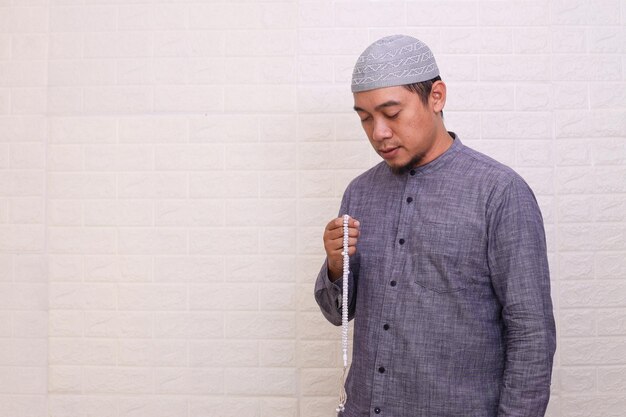 Religious Asian muslim man praying with eyes closed and dzikr with prayer beads isolated on white ba