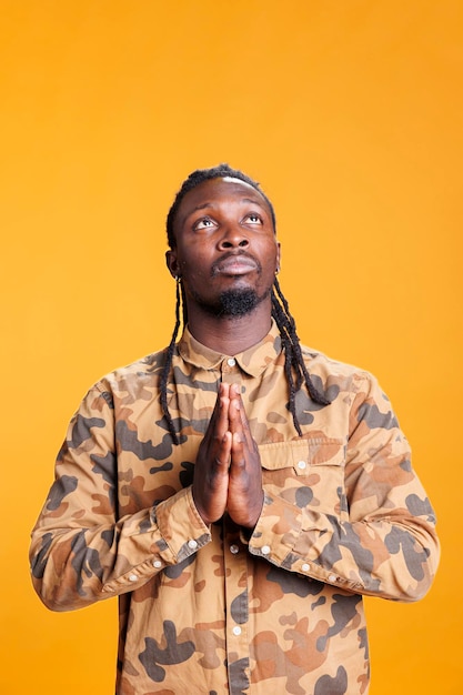 Religious person holding palms together in pray symbol in studio, showing spiritual gesture over yellow background. Faithful african american man praying to jesus for peace and forgiveness.