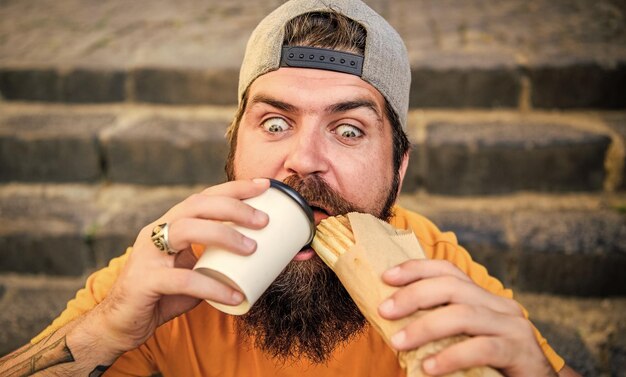 Relish the moment Hipster drinking coffee with hotdog snack on stairs outdoor Bearded man dining with unhealthy sandwich and beverage snack Hungry guy enjoying snack on street Snack and food