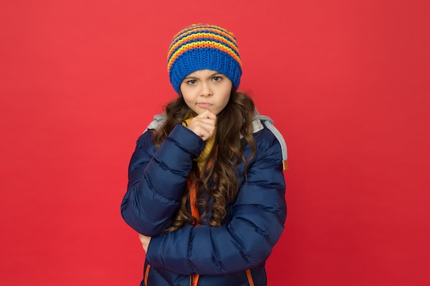 Riluttante a fare qualcosa. bambino dubbioso premuroso. bambina indossare abiti invernali sfondo rosso. concetto di infanzia. cappello lavorato a maglia capelli lunghi ragazza emotiva. stile casual da ragazza teenager. emozioni e umore.