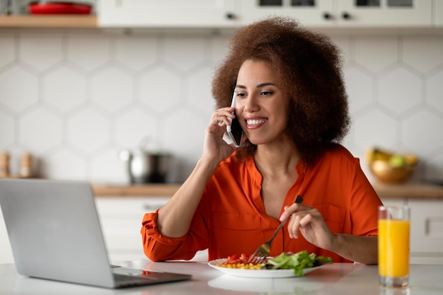 Photo remote business black woman using cellphone and working on laptop in kitchen