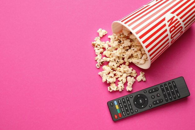 Remote control and cup of popcorn on pink background flat lay Space for text