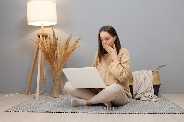 Foto lavoro a distanza opportunità freelance online donna caucasica scioccata che indossa un maglione beige che lavora al computer che ha problemi a salvare le informazioni mentre è seduta sul pavimento in una stanza luminosa