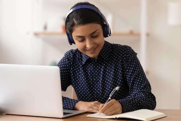 Remote student focused young indian lady wearing headphones
practice in distant learning of english language write test before
pc screen hindu woman in earphones listen to webinar online take
notes