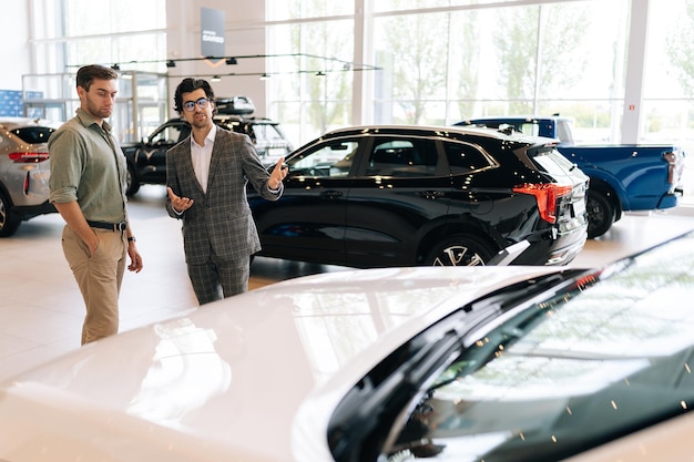 Photo remote view to professional car dealer in business suit telling pensive male buyer about luxurious