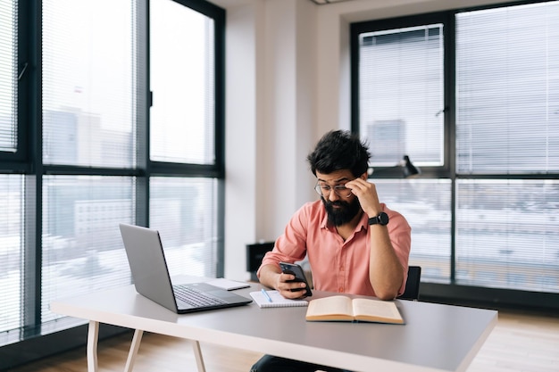 Remote view of puzzled Indian freelancer male in glasses using smartphone sitting at office desk with laptop and paper documents by window Relaxed bearded businessman using mobile phone typing email