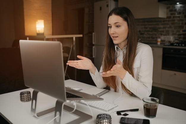 Remote work. A caucasian woman with headphones working remotely online on her laptop. A girl actively discussing business with her colleagues through a video call at her cozy home workplace.