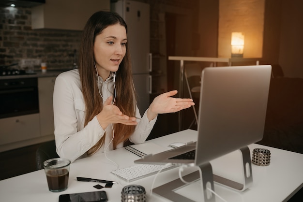 Remote work. A caucasian woman with headphones working remotely online on her laptop. A girl actively discussing business with her colleagues through a video call at her cozy home workplace.