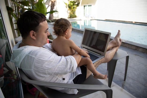 Lavoro a distanza. uomo con un bambino in braccio con un laptop seduto a bordo piscina in un paese tropicale.