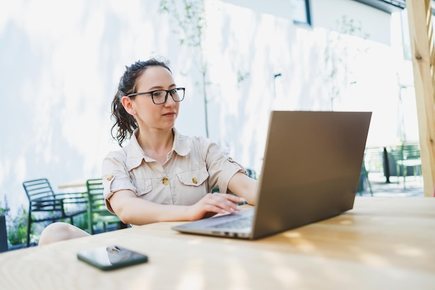 원격 작업: 카페의 야외 테라스에 앉아 커피와 전화로 온라인에서 일하는 편안한 여성 여성 프리랜서 여성은 여름 카페에서 노트북에 앉아 온라인에서 원격으로 일합니다.
