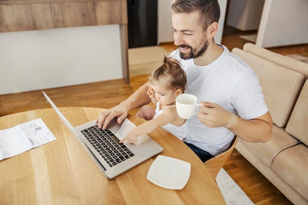 A remote worker is holding his infant in a lap and working on a laptop while she is helping him