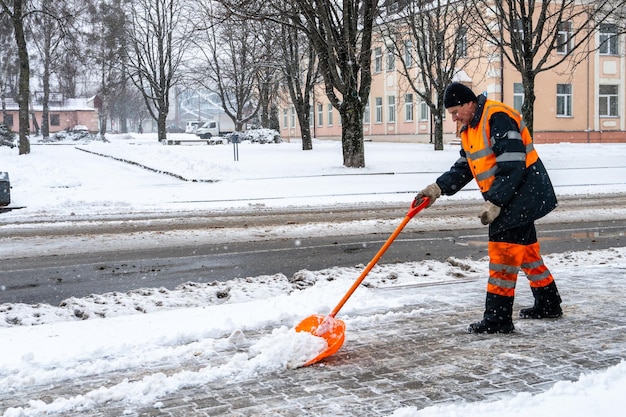 Фото Уборка снега с тротуара после метели дорожный рабочий с лопатой в руках и в специальной одежде очищает тротуар и дорогу от снега метель и ураган в городе