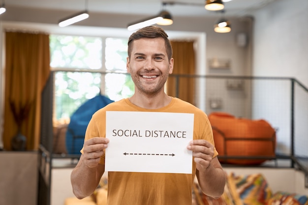 Reopening office safely young happy man showing paper with text social distance at camera and