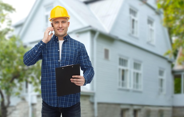 repair, building, construction, real estate and maintenance concept - smiling man or builder in helmet with clipboard calling on smartphone over living house background