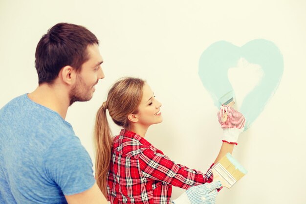 концепция ремонта, строительства и дома - улыбающаяся пара рисует маленькое сердце на стене дома