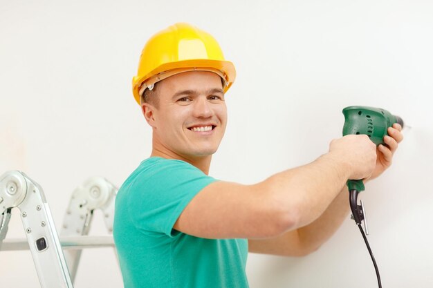 repair, building and home concept - smiling man in yellow protective helmet with electric drill making hole in wall