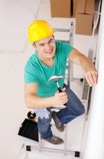 修理、建物、家の改修のコンセプト-壁に釘を打つ黄色の保護ヘルメットの笑顔の男