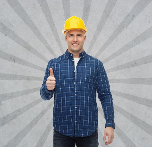 Photo repair, construction, building, people and maintenance concept - smiling male builder or manual worker in helmet showing thumbs up over gray burst rays background